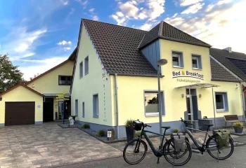 Außenansicht der gemütlichen Pension Gästehaus Legden mit zwei Touren-E-Bikes im Vordergrund vor dem gelben Gebäude unter blauem Himmel.