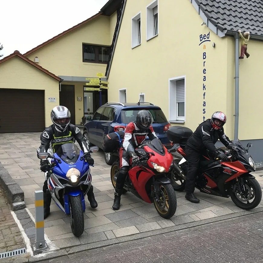Biker willkommen! Unsere Pension in Legden ist der ideale Boxenstopp für Ihre Motorradtour durch das fahrfreundliche Münsterland. Der befestigte und großzügige Hof bietet Gruppen eine bequeme Stellfläche für ihre Maschinen direkt an der Unterkunft – perfekt für einen entspannten Zwischenstopp oder als Basis für Tagestouren. Drei Motorradfahrer auf Sportmotorrädern in der gepflasterten Einfahrt vor der gelben Außenfassade der Pension in Legden.